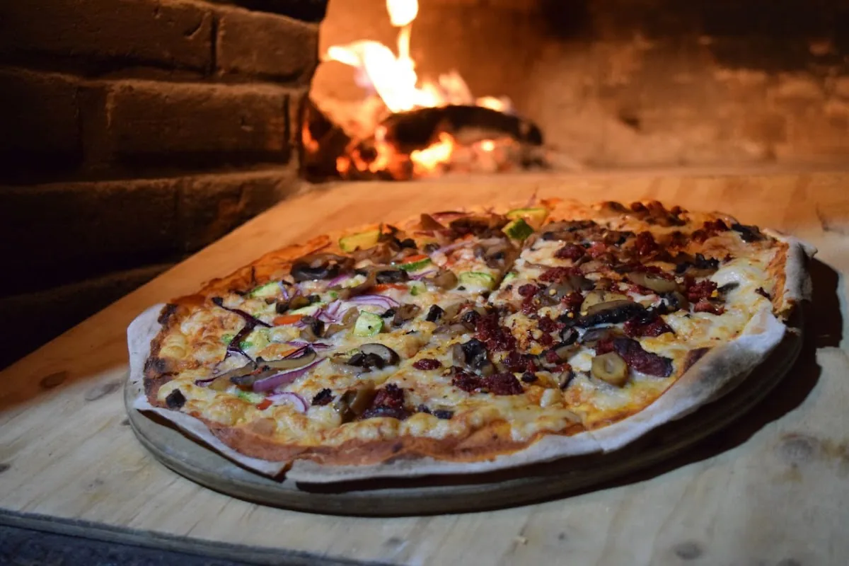 Pizza recién salida sobre tabla de madera, con el fuego del horno de leña atrás