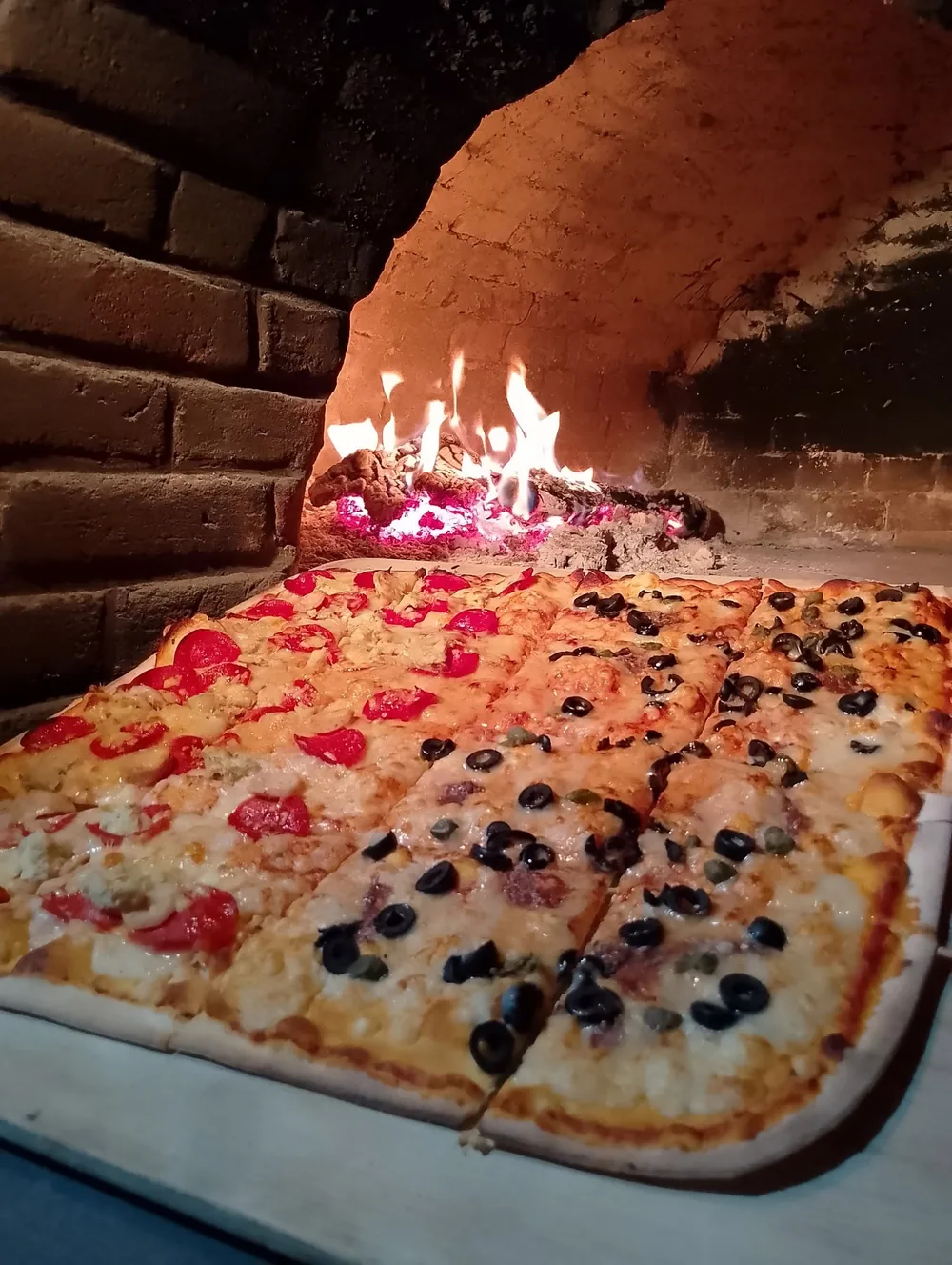 Pizza rectangular de pepperoni y aceituna negra frente al fuego del horno de leña